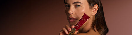 Woman with glowing skin holding Tropic Skincare Eye Lift Exosome Midnight Balm close to her face against a warm brown background, showcasing the product’s luxurious red and gold packaging.